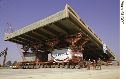 A self-propelled modular transporter (SPMT) moves the prefabricated superstructure of the 4500 South bridge in Salt Lake City, UT. Three workers are visible in front of the SPMT.