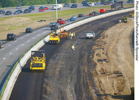 Highway lanes closed off for construction on the right side of a highway. Construction equipment and workers are visible. Traffic is traveling on the left side of the highway.