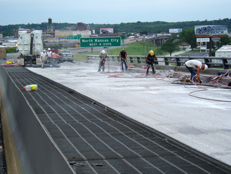 Six construction workers are shown working on an overpass within a highway work zone. A sign for North Kansas City is seen in the background.