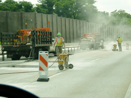 Roadside view of two workers operating within a work zone on the side of a highway. Trucks, dust, and machinery surround the workers.