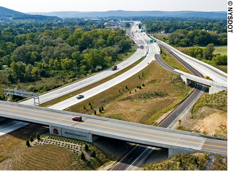 An aerial view of a highway interchange. Two overpasses cross over multi-lane highways. Vehicles are traveling on the highways. Wooded areas can be seen in the background.