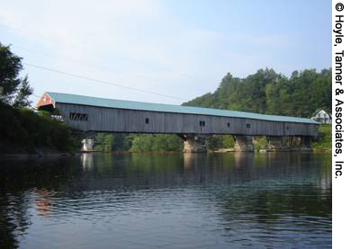 A side profile view of the Bath Village Covered Bridge in Bath, NH. The wooden bridge is shown stretching out across the water.