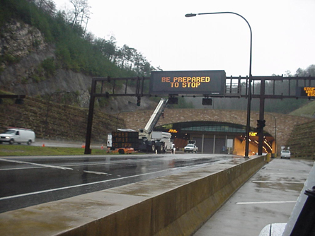 A view of U.S. Route 25E approaching the entrance to the Cumberland Gap Tunnel. An electronic overhead sign has the message "Be Prepared to Stop." A truck with an aerial lift is in front of the sign.