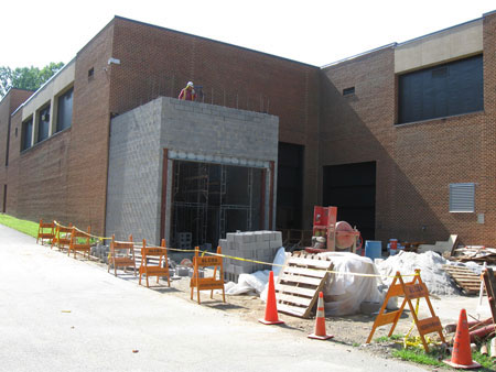 A two-story masonry building is shown, with construction materials and equipment outside and a worker in hardhat observing from atop a partially constructed concrete block addition.