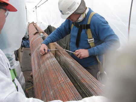 Two bridge inspectors in hardhats are examining the strands of a large suspension cable. The inspection area is enclosed in plastic.