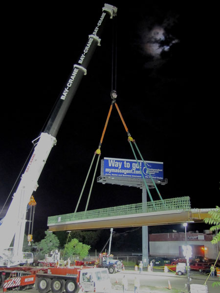 A night-time view of work at the Fast 14 project site. A crane is lifting part of the bridge superstructure. In the background is a billboard that reads "Way to go!"