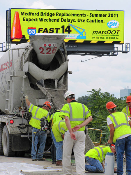 Five workers are in the foreground in front of a concrete truck. In the background is a billboard that reads "Medford Bridge Replacements-Summer 2011. Expect Weekend Delays. Use Caution." The Fast 14 project logo and MassDOT logo are on the billboard, along with a logo for 511.