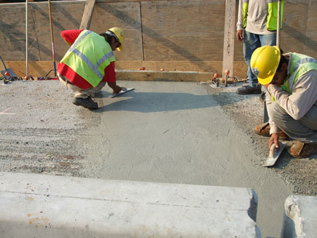 Two workers smooth concrete over a closed bridge deck joint.