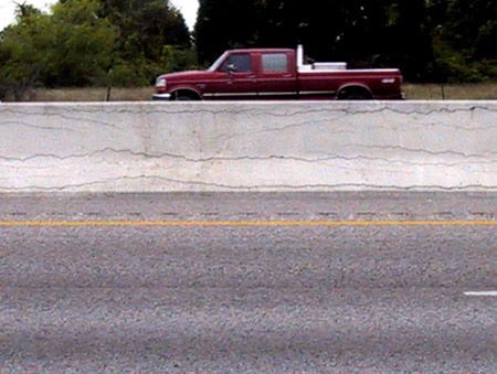 A view of a concrete highway barrier that is exhibiting cracking caused by alkali-silica reaction (ASR). On the other side of the barrier is a pick-up truck.