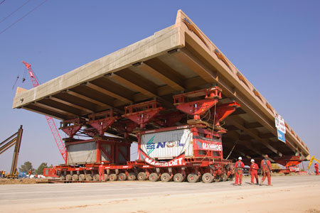 A view at a bridge construction site of a prefabricated multi-lane bridge section with eight visible girders being moved into place using a self propelled modular transporter (SPMT). This extra-wide multi-wheeled apparatus is positioned transverse under the bridge section. Workmen in safety gear are looking on. A crane and other equipment are visible in the background.
