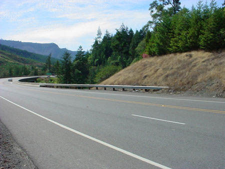 A close-up view of a highway in Oregon with a guardrail in the background. A car is traveling in the distance.