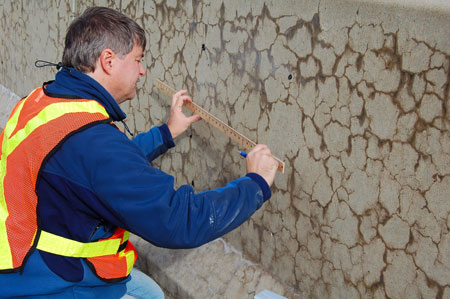 A highway worker wearing a safety vest uses a ruler to examine a concrete median barrier that is affected by alkali-silica reactivity. The barrier has cracks in the concrete.