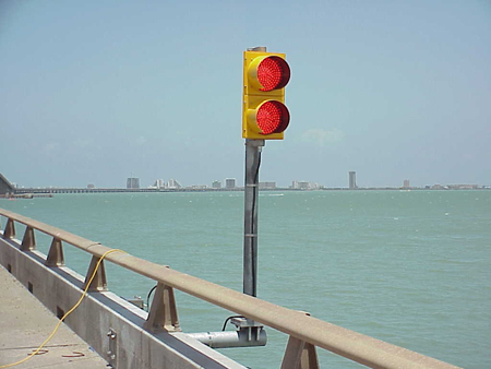 A close-up view of two traffic signals flashing red on the Queen Isabella Causeway bridge in Texas. The traffic signals are mounted to the side of the bridge. In the background is the Gulf Coast Intracoastal Waterway.