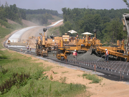 A view of a continuously reinforced concrete pavement under construction. Paving equipment and workers are visible. In the background are wooded areas.