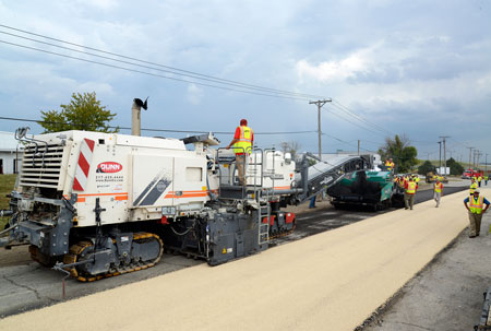 Workers operate a paver. Six workers wearing safety vests are visible.