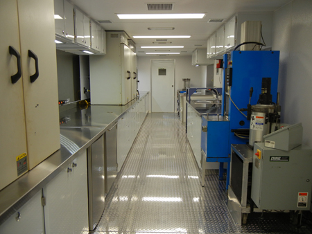 A view of the length of the interior of FHWA's Mobile Asphalt Testing Lab. Equipment, counters, and cabinets line the walls of the tractor-trailer truck. To the right in the foreground is the Asphalt Mixture Performance Tester, which can be used to measure the dynamic modulus, flow number, and fatigue properties of an asphalt mix.