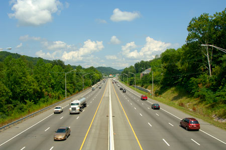 A head-on view of a divided highway. Three lanes of traffic are traveling in both directions. Both cars and trucks are visible. Trees line the shoulders of the highway.