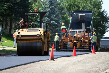 A view of an aspalt road being paved. Four workers wearing safety vests and hard hats are visible. In the foreground is a roller. A dump truck is behind the roller. Three traffic cones are placed on the edge of the road being paved.