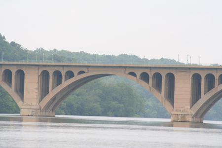 Partial side view of a large, masonry, open-spandrel, multiple-arch bridge over still water with guardrail and streetlights atop bridge. A forested hillside is in the background.