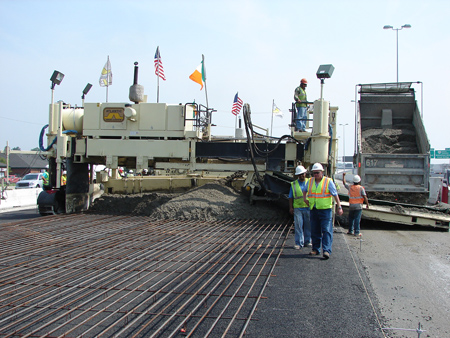 A close-up view of a paving operation for a continuously reinforced concrete pavement. Continuous steel bars are being placed on the roadway. Four construction workers wearing hard hats and safety vests are visible. Paving equipment and a dumptruck are in the background.