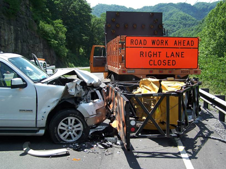 A close-up side view of a car that has hit a work zone barrier on a roadway. The hood of the car is smashed in and debris is on the ground. Behind the barrier is a dump truck with a sign affixed to the rear of the truck that reads "Road Work Ahead. Right Lane Closed." (Photo Credit: © VDOT).