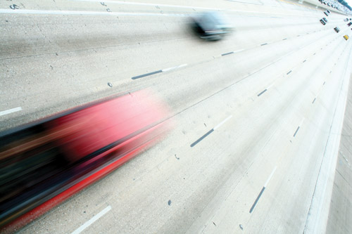 Photograph taken from an overpass showing two cars with motion blurs speeding by on a multi-lane highway below.