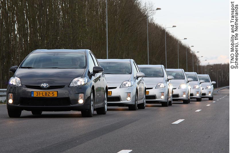 Daytime photo of a platoon of six cars. A black Toyota Prius leads a platoon made up of five silver Priuses. The oncoming cars are driving closely together (less than a car length apart) on a two-lane highway; there are no other cars on the road. It is daytime, but all of the cars have their headlights on. The cars all have Dutch license plates.