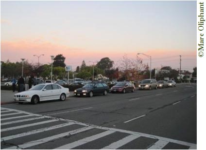Six cars are lined up along multi-lane street waiting for passengers at a train station parking lot. It appears to be early morning: the sun is rising in the background and there is no traffic on the street aside for the queuing cars. Some of the cars have their lights on. In front of the queuing cars a crosswalk is painted on the street. A full parking lot lined with trees and street lights can be seen behind the line of cars.  A few people are milling around the first car in the line.