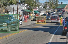 Displayed urban street scene with cars moving in two directions and pedestrians about to cross mid-block. Pedestrians and approaching cars are highlighted with colored boxes in the display.