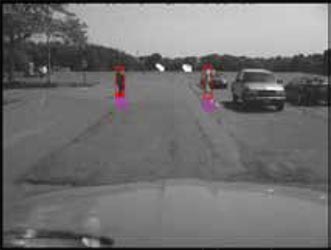 Display camera view from inside car showing car hood, parked cars ahead, and two pedestrians crossing a street. Bright pink rectangles are superimposed over the pedestrians in the display.