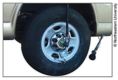Photo of a microphone mounted on the underside of a vehicle and pointing at a tire. 