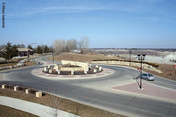 A roundabout in a rural area with roadways entering from the top, bottom, and left sides of the photo. A raised brickwork median separates traffic lanes where the roadways enter the circle. On the left side of the roadway is a sidewalk, and on the right side there is a low stone wall with shrubbery behind it. In the center of the circle is a landscaped area containing a low stone wall in the shape of a circle dissected by a taller stone wall in the direction of the roadway that enters and leaves the circle; and at right angles, a lower wall pointing to the roadway that completes the junction. Just outside this structure are white cube-shaped markers with black arrows pointing clockwise (the direction of traffic flow). Outside this central area is a wide patterned brickwork circle incorporating white arrow heads that also point counterclockwise. A car is entering the circle in the foreground. On the other side of the circle a pedestrian is crossing the road. Four sets of streetlights are visible. Houses and trees appear in the distance.
