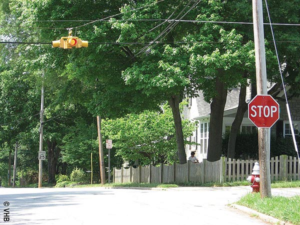 View of an intersection in a residential area, with “STOP” sign and fire hydrant on the right, a yellow traffic light suspended on cables above the center of the intersection, a 25 mi/h speed sign in the distance, and a home on one corner.