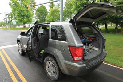 View from rear and side of a gray, SUV-type, test vehicle parked at a demonstration intersection with driver's side rear door and hatchback door open and testing equipment visible inside. Testing sensors are also mounted on the tires.