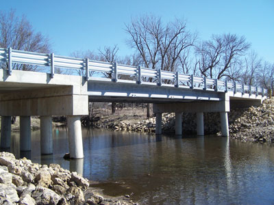 View from ground-level of a short bridge mounted on 4-legged pi-girders, one near each shore of a narrow river.