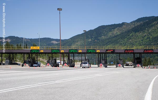 View of a highway toll plaza with seven toll stations and a few vehicles visible. Hills are in the background.