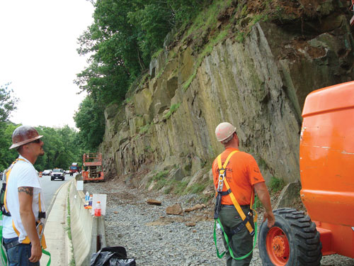 View of construction at the side of a traveled roadway. Two workers in safety helmets are examining a sheer rock face separated from the roadway by a Jersey barrier and several feet of flat, graveled space. Small pieces of fallen rock and two pieces of equipment are in the picture.