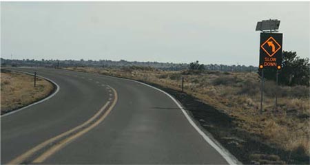 Photo of a curved two-lane rural road with an electronic roadway sign with a curved arrow to the left and text stating slow down
