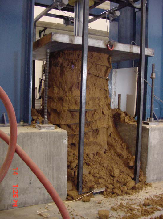 This photo shows the test specimen from figure 14 following compression testing. The mass is no longer a solid cylinder. The outside is ragged and crumbling, and soil has begun to pile up at the bottom of the mass. 