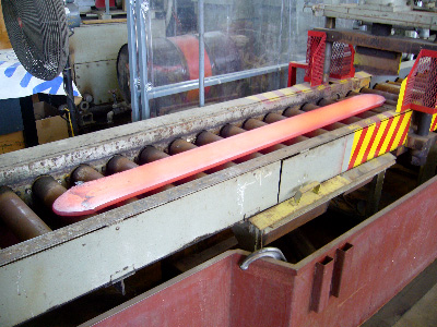 In this photo, the same piece of steel from figure 8 is shown a few minutes after it was hot rolled from 5 to 0.5625 inches (127 to 14.3 mm) thick. The photo shows the delivery side of the reversing mill, and the steel is a dull orange. The steel has elongated from about 11 to 100 inches (280 to 2,540 mm).