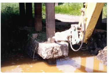 Shearing of pile of bent 2. This image shows the south pile/column of bent 2 of the Trinity River Relief Bridge being sheared and broken using a vibrating breaker point on a backhoe. The water level in the excavation is near the bottom of the strip footing.
