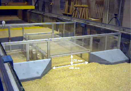 Figure 2. Photo. Rectangular culvert. The photo shows the model of the rectangular bottomless culvert used in Phase II of the bottomless scour experiments. The model, made mostly of Plexiglas, is centered in the flume. The model’s entrance is to the front. A wingwall is on each side of the entrance. The model’s back is to the left rear. The bottom of the model is sand. The dimensions of the entrance to the culvert are superimposed on the photo. The entrance is 150 millimeters high and 600 millimeters wide. Wingwall models are on each side of the entrance.