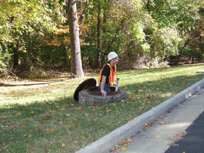 Figure 1. Photo. A typical access hole. This picture, taken from the street, shows a man with a hard hat and reflective vest climbing out of an open manhole (or access hole) via a ladder. The manhole is located in a grassy strip between the street and a wooded area, and the manhole cover is leaning against the concrete exterior of the manhole on the far left side.