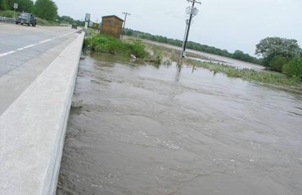 This photo is taken looking down the length of a bridge with only the upstream edge of the bridge depicted in the left of the photo. The water at the right of the photo is very high, and it appears to rise to just a few feet below the road deck, but it does not overtop the bridge.