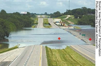 This photo looks down a divided highway with two lanes on each side. The river crosses over the two sides of the highway at the center of the photo with water flow going from right to left. The upstream edge of the bridge deck appears to have guard rails just above the water, but otherwise, both spans are completely submerged.