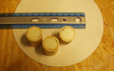 This photo shows three pipes that contain a tag that were then filled with epoxy and cured sitting on a circular piece of paper. A ruler placed above the pipes shows scale. The pipes are 3/4 inches in diameter. Air voids between the inner wall of the pipe and the epoxy can be seen in all three pipes.