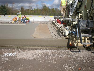 This photo shows the finished paved concrete road, which is 13 inches thick. At the right of the photo is the slip form paver that has just finished paving. In the background, three workers are checking the air content of the concrete, the slump and finishing the concrete with a screed.