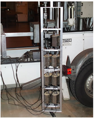 Figure 124. Photo. Seismometer stand with sensors attached. This photo shows seven seismometers attached to the bottom positions in a two-column seismometer stand. The 
stands are in a room positioned vertically on the ground, and the frame of the falling weight deflectometer is seen in the background.
 