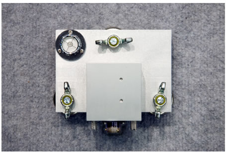 Figure 13. Photo. Accelerometer box on calibration platter. This photo shows a top view of an aluminum accelerometer box attached to an aluminum calibration platter. The box is attached to the front of the platter by two knurled thumb screws. Three wing nuts attached to feet are used to level the platter. A bubble level on the back left corner of the platter is used to confirm that the platter is level.