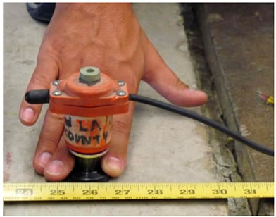 Figure 39. Photo. Locating a place to install the ball-joint floor anchor. This photo shows a hand manually holding a red JILS geophone 4 inches (100 mm) from the edge of a concrete test slab. A ruler in the foreground shows the distance.